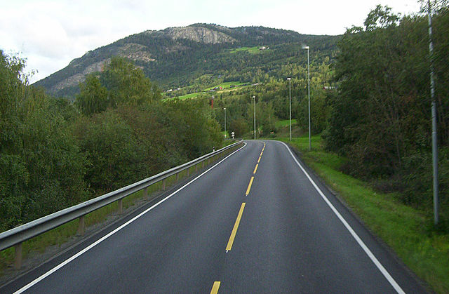 Picture of a straight in an open road leading up to a left hand bend in the distance with a lush green setting of trees lining either side with mountains in the background